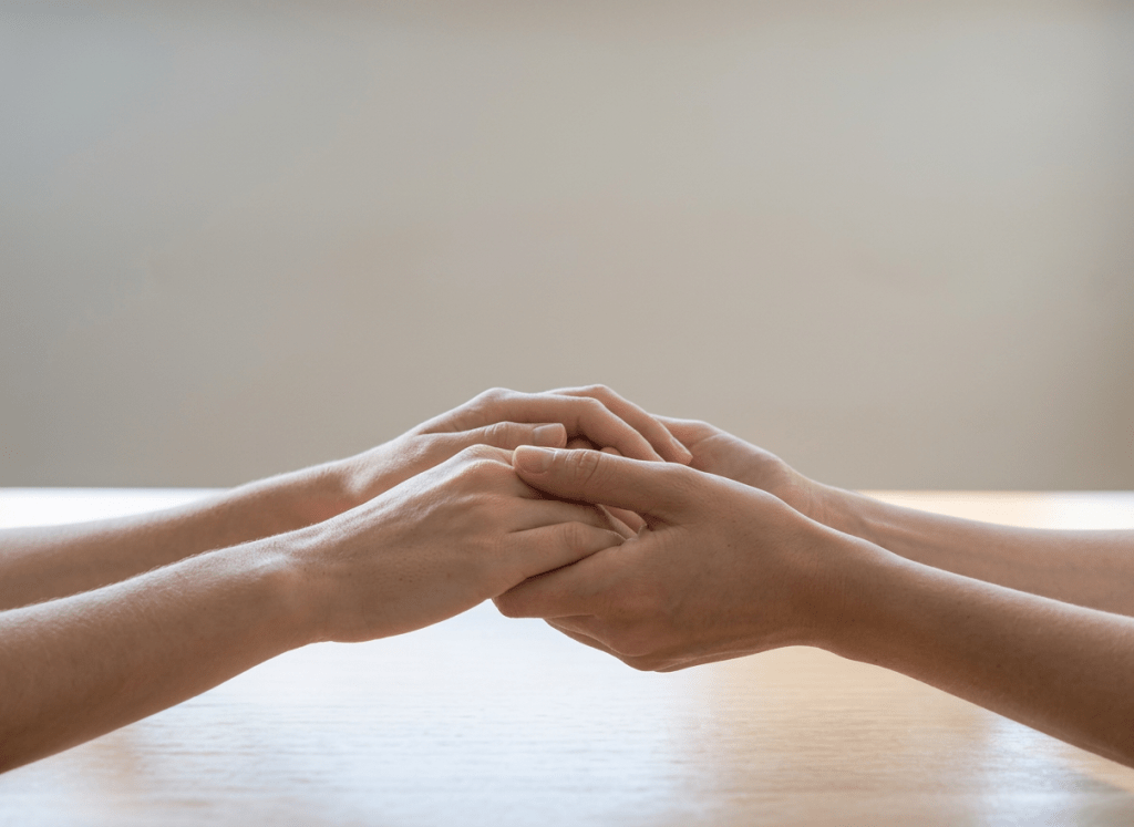 Two pairs of hands gently holding each other on a table near an open window